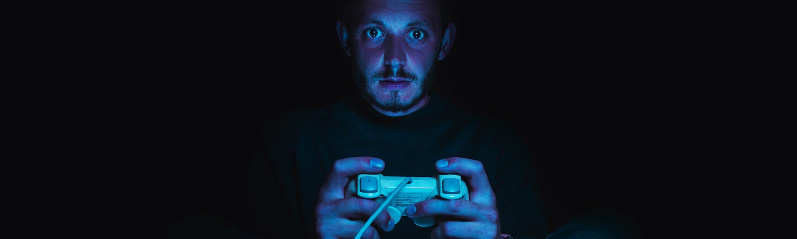 A man sits in the dark holding a video game controller. His look is mesmerized by the blue light of the game.