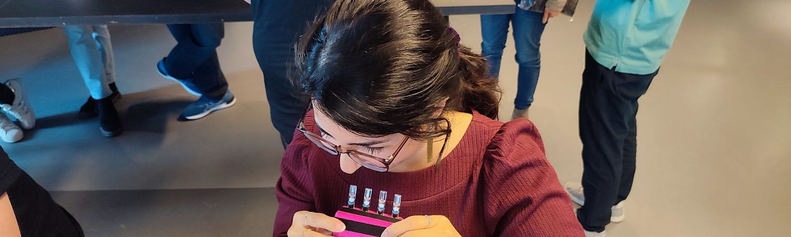 A woman with dark hair in a ponytail is sitting in front of a laptop and holding a pink 3D-printed sip & puff device whilst reading a sheet of commands.