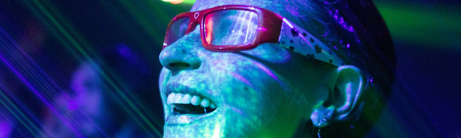 This image depicts a person at what seems to be a party or event where UV lighting is in use, as indicated by the vibrant greenish glow on their face and clothing. The person is wearing red-framed glasses and earrings, and appears to be having a joyful moment, with their mouth open as if they are laughing or cheering. The background is blurred, but lights and possibly other people can be seen, suggesting a lively atmosphere. The visible laser lights add to the festive ambiance of the setting.