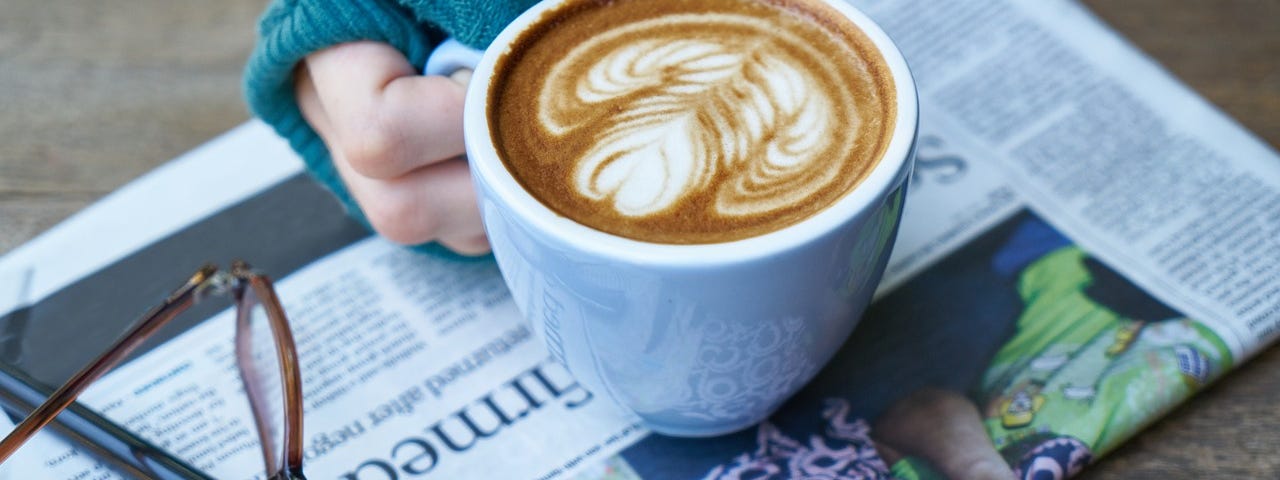 A hand holding a cup of coffee over a newspaper with a pair of glasses and a pen.