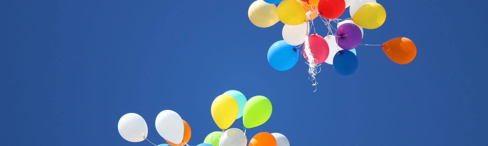 Two brightly coloured bunches of balloons released into a clear blue sky.