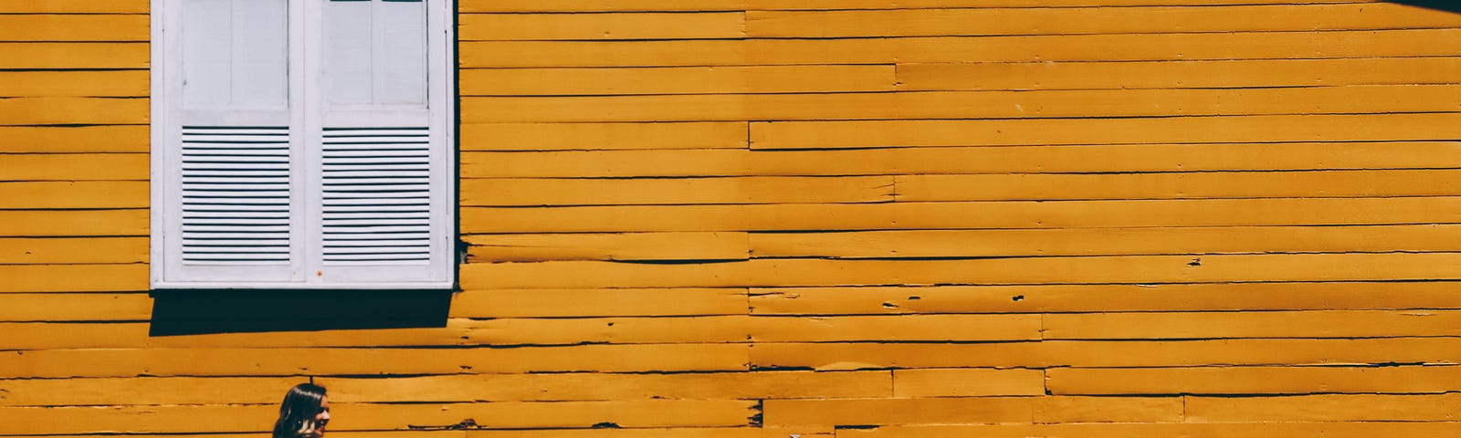 a woman in a white dress against a yellow wall made of wooden planks with a white window above her