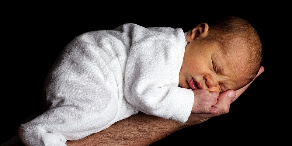 Newborn baby sleeping on father’s forearm and palm. White pj’s contrast with Black background.