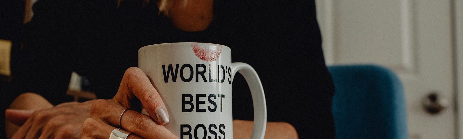 A hand holding a mug that reads: “World’s Best Boss”. The hand has long fingernails with nail polish, and the mug has a lipstick mark on it.