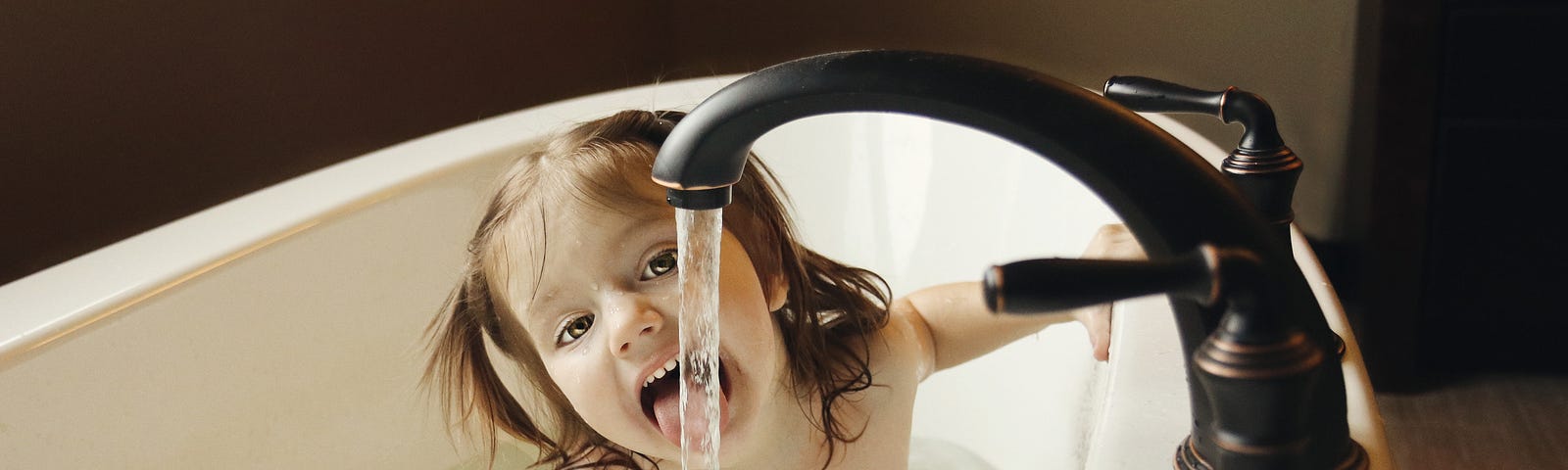 A young girl in a tub, trying to drink water from the faucet