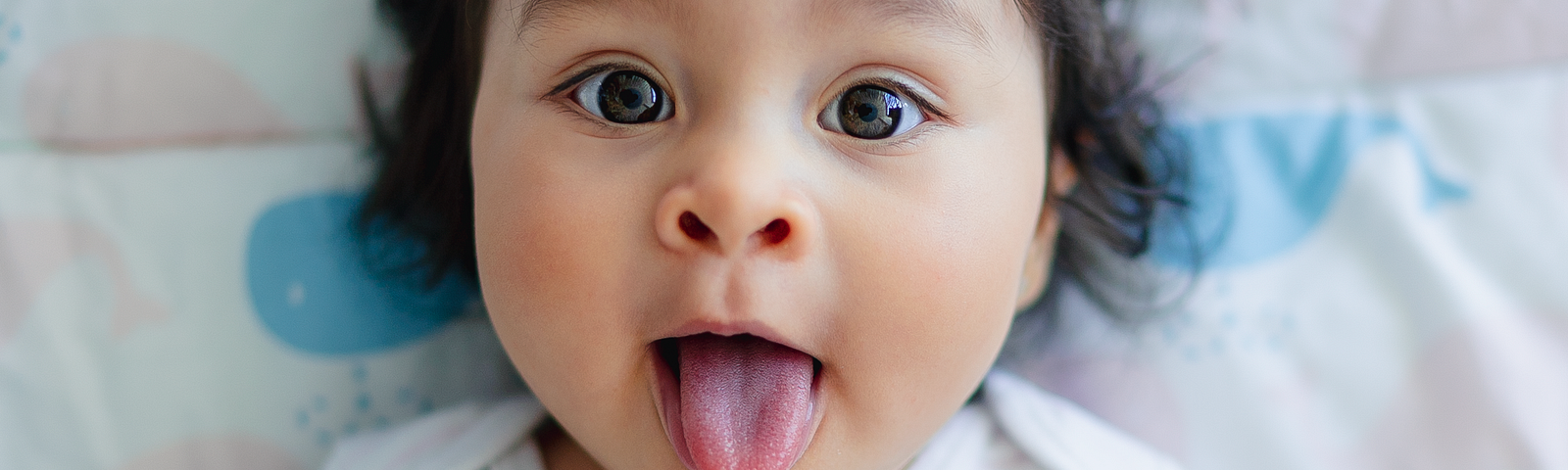 On a baby’s little shirt is written “One of a Kind.” The infant has a round face, dark hair, and sticks our her tongue at the photographer.