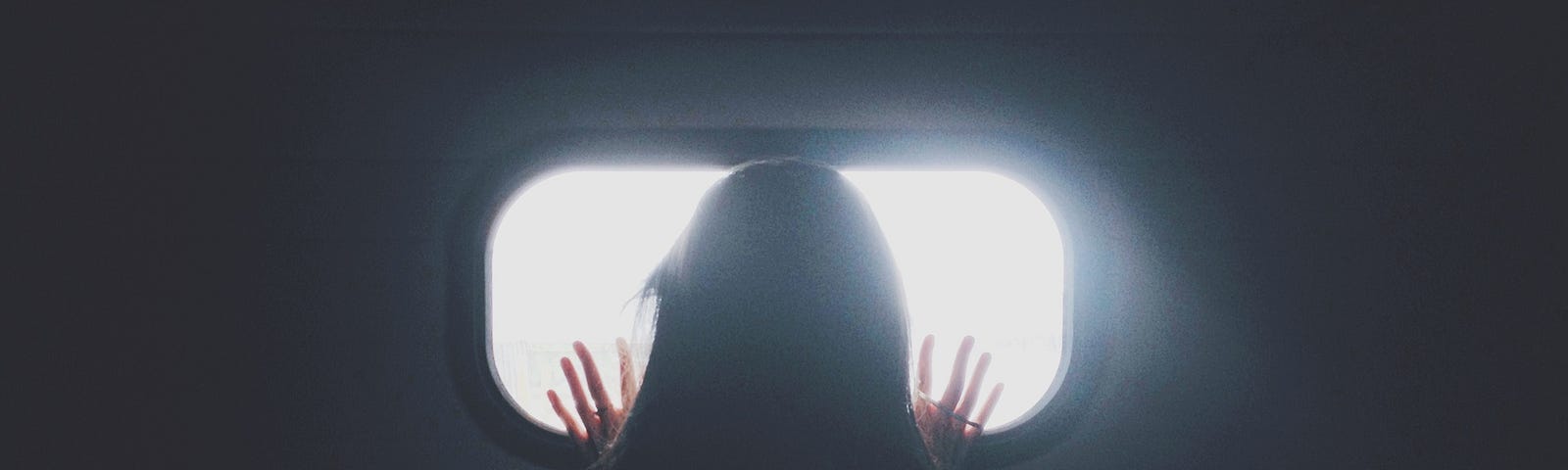 A woman with her back turned, looking out a window in a dark room.