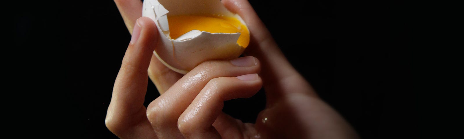 A shattered egg drips yolk into a hand holding the shell.