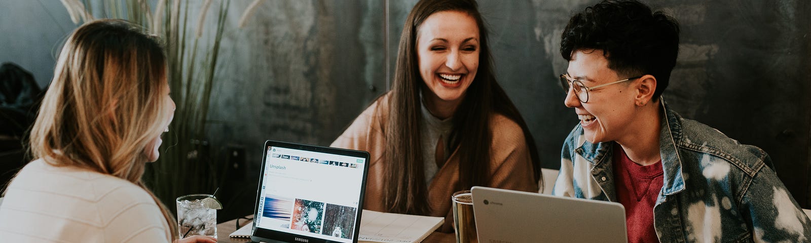 A group of three women sit at a wood table with laptops. They are laughing and working together.