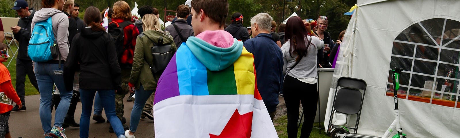 A young person wears a Pride flag across his back, embossed with the Canadian red maple leaf.