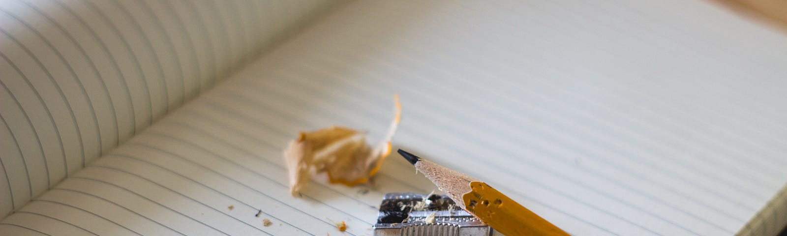 a freshly sharpened pencil on a blank lined notebook with a small pencil sharpener and pencil shavings on the page.