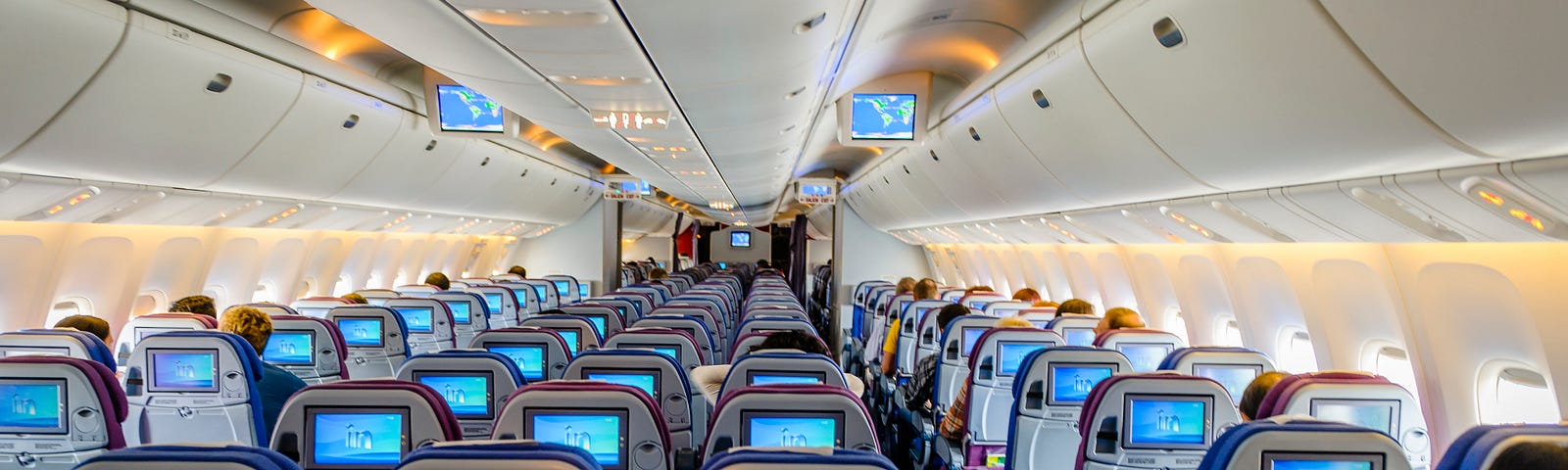 An aircraft cabin with rows of seats, some of which have people sitting in them, watching the T.V. screen on the back of the seat in front.