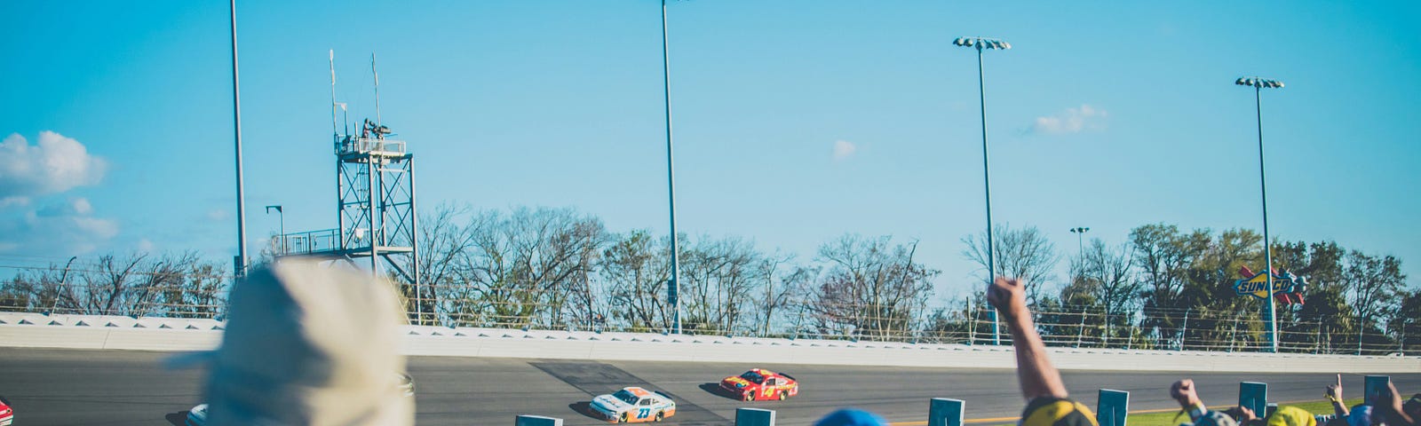 Race fans at a speedway cheering on race cars as they round the track coming out of a turn into the straightaway