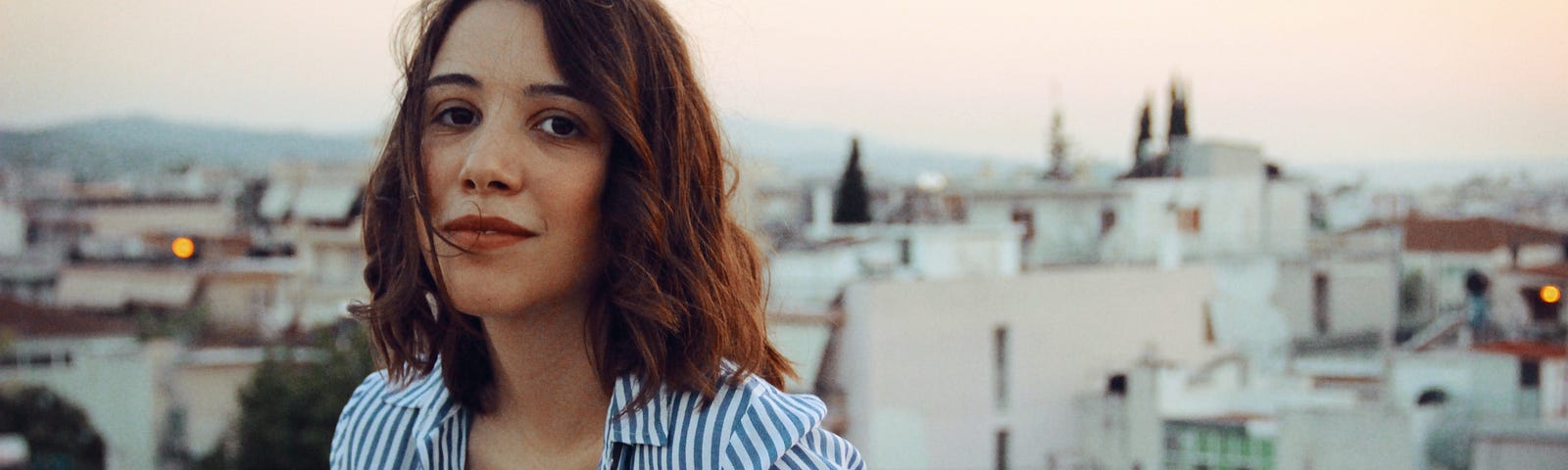 A mid-shot of a young Hispanic woman standing with her back leaning against a balcony. The white villas and pale pink sky behind her suggest she is in a hot climate, maybe Spain. She’s wearing an open stripy white and blue shirt and her mid-length brown hair blow gently as she smiles knowingly at the camera. I Love Your Honesty, Not Your Sales Tactics - Food for thought for all online entrepreneurs and business owners when pitching your digital product. This will actually make me want to buy it.