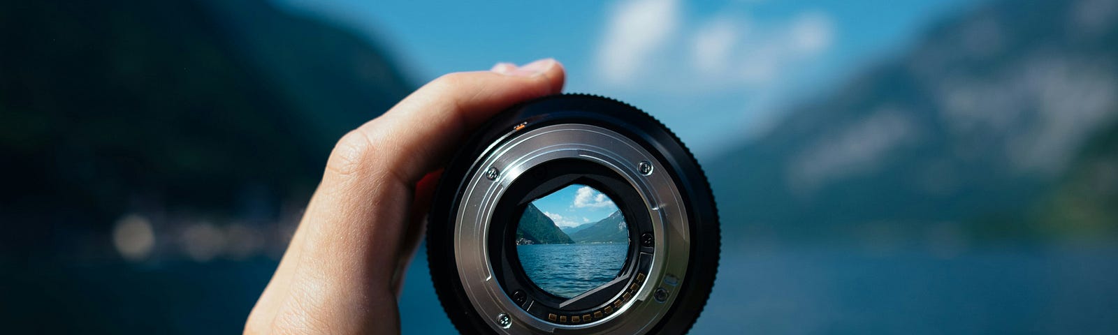 A hand grips a camera lens without the body of the camera attached. Through the lens you get a much more focused view of a lake and the mountains on the horizon.