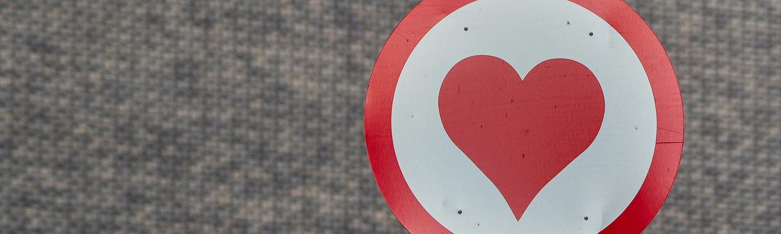 A traffic sign with a red heart in the center posted on a metal pole, with a grey textured background.