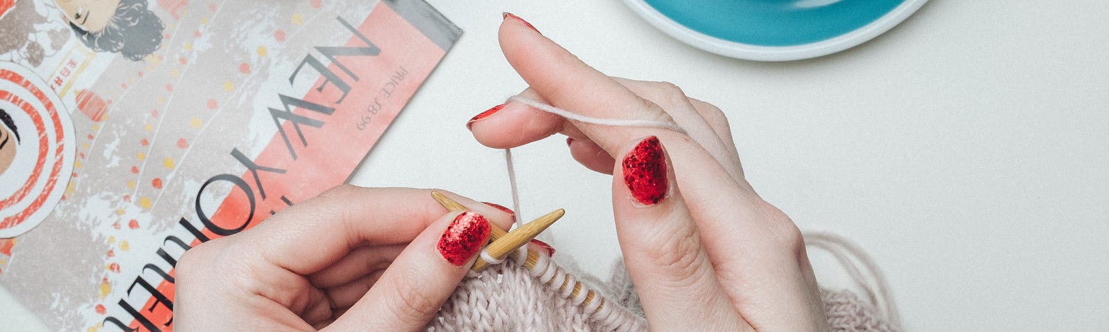 Picture of someone’s hands knitting on a project over a white desk, with a cup of coffee and a magazine nearby.