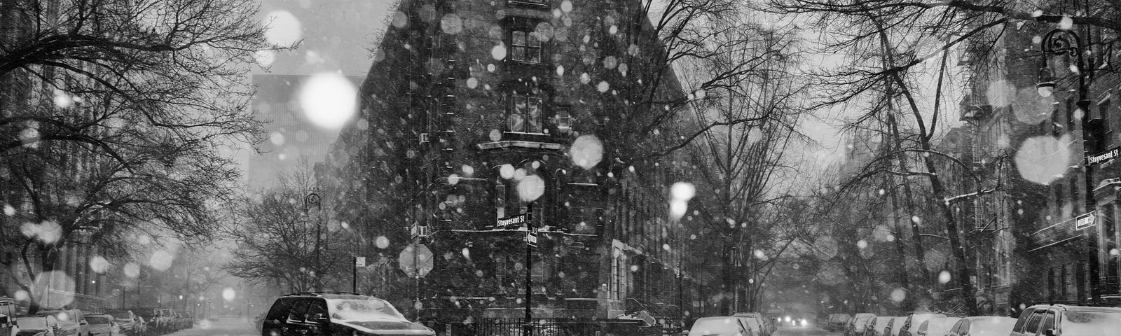 Intersection of city streets during heavy snowfall. Tall buildings and trees, cars parked, one car driving, cinders on street