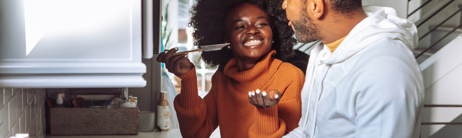 A Black woman in a kitchen with her partner. She is holding up a spoon and grinning widely, as her partner chops up some tomatoes.