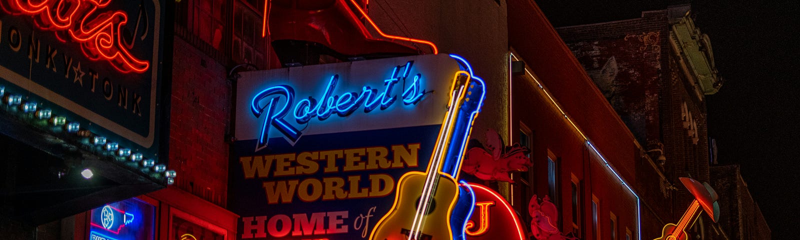 Nighttime street scene on Broadway in Downtown Nashville. Neon signs for Robert’s Western World, Jack’s BarBQue, and The Stage are in the background.