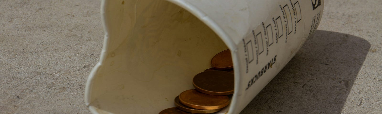 A dirty paper cup on the ground with coins inside.