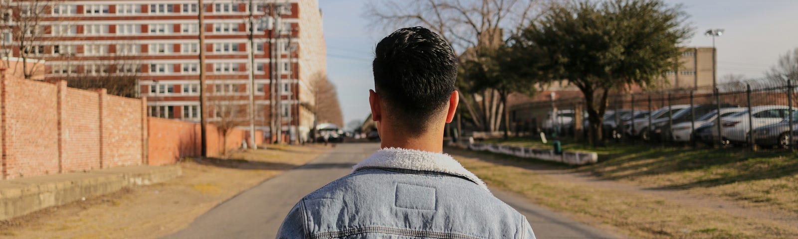 A man with short hair wearing a denim jacket. The man is facing away from the camera looking at an empty road ahead of him. There are buildings and a car park on the side of the road.