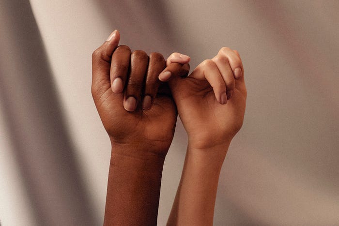 Two hands — one white and one black — reaching up and holding pinkie fingers.
