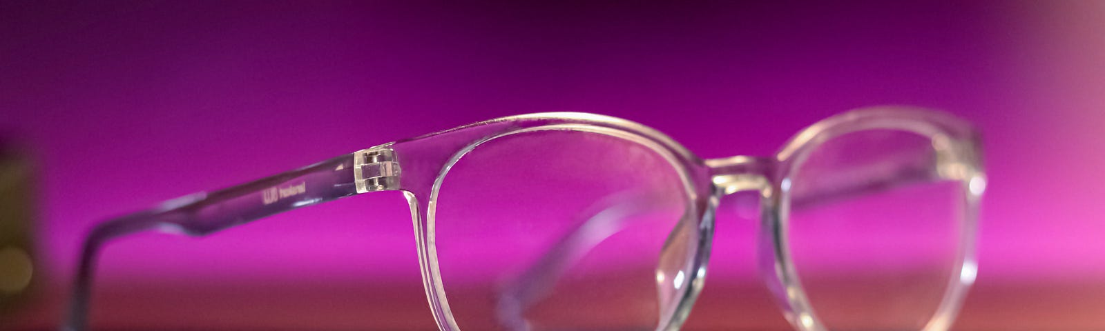 Clear frame eye-glasses looking, at an angle, to viewer — sitting with a mauve background.