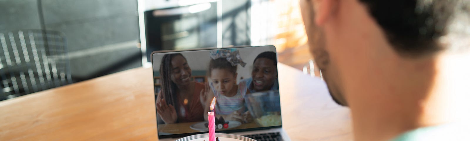 A photograph of a person holding a cupcake with a candle up to a laptop screen. The laptop has a video call with two adult and a young child, who is leaning forward and blowing towards where the candle is.