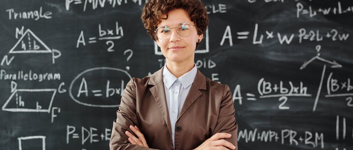 A woman with short brown hair, in a brown jacket, standing in front of a chalk board with formulas on it with her arms crossed.