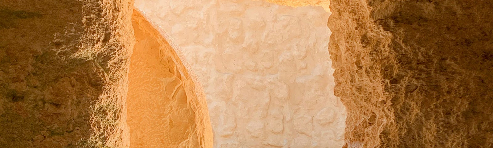 This image shows an ancient tomb entrance carved into a sandstone wall, creating a tunnel-like pathway that leads to a sunlit area with visible stone rubble. The warm lighting enhances the texture of the rocky surfaces, highlighting natural earth tones ranging from deep orange to sandy beige. The passage is narrow, with rough and uneven walls that suggest a natural or minimally modified state.