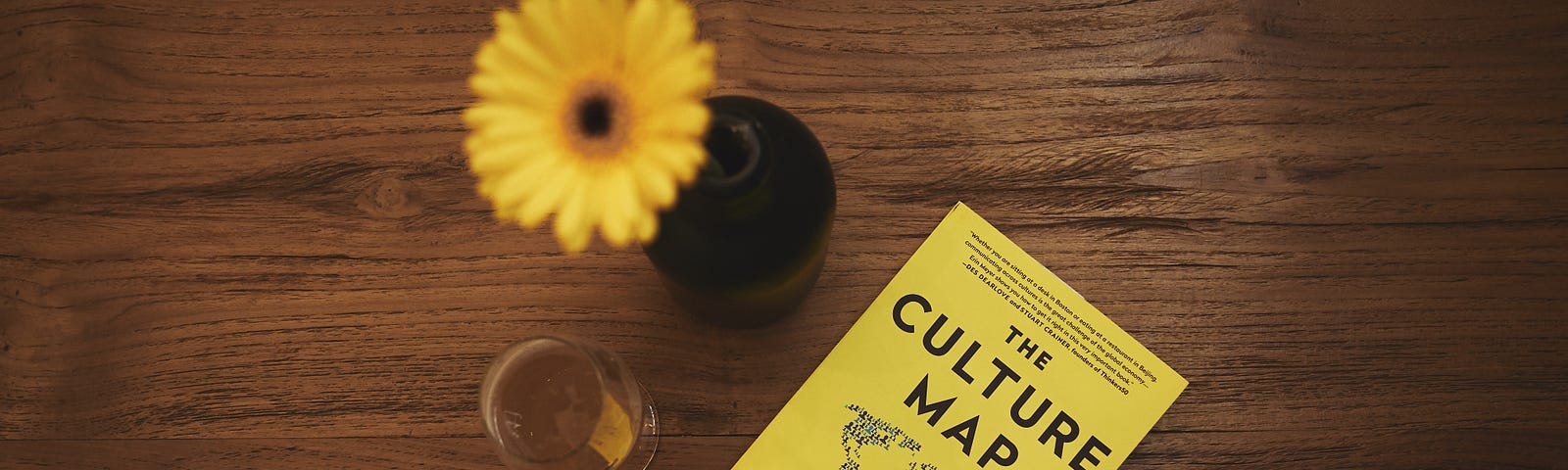 A yellow daisy in vase next to white wine glass and yellow book titled, “The Culture Map” by Erin Meyer on a brown table.