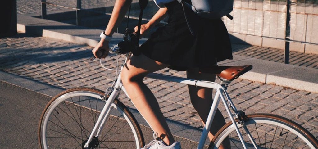 Mulher branca de saia preta, tênis e mochila, em uma bicicleta branca com banco de couro.