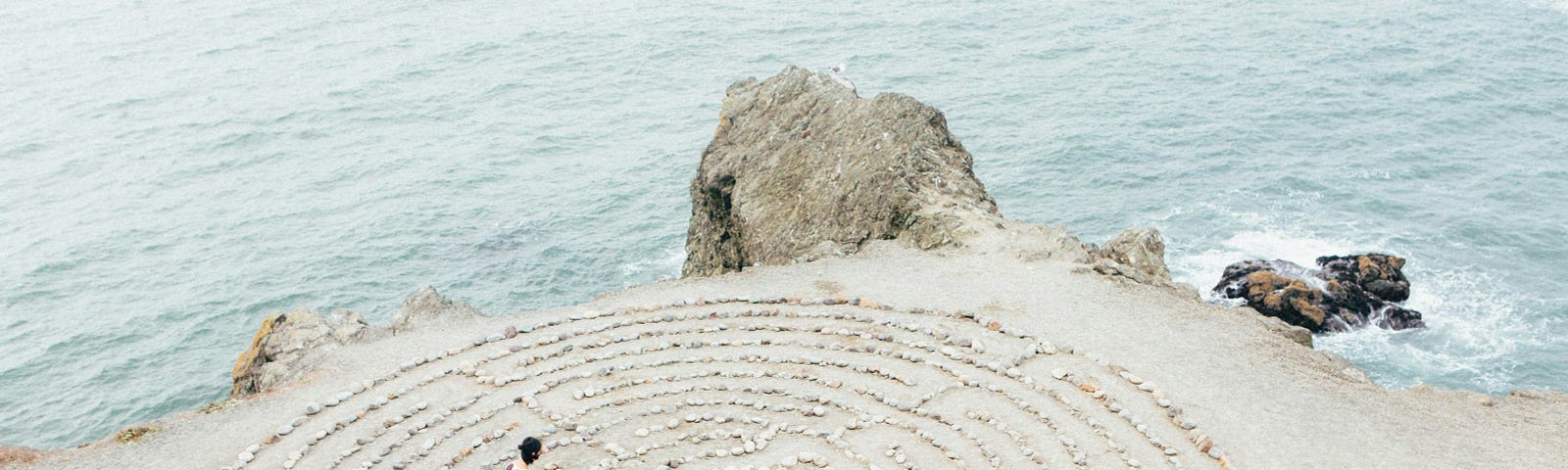 An overhead shot of a person walking through a labyrinth of shells on a sandy beach straddling a rocky shoreline