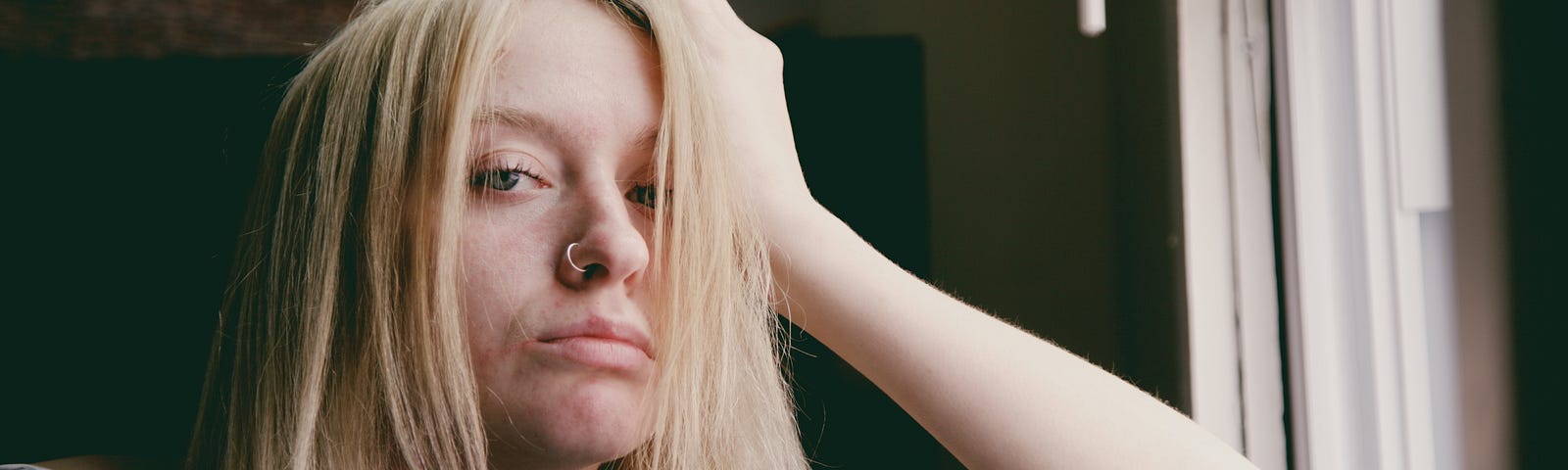 A tired woman with blonde hair rests her arm on a windowsill and her head in her hand.