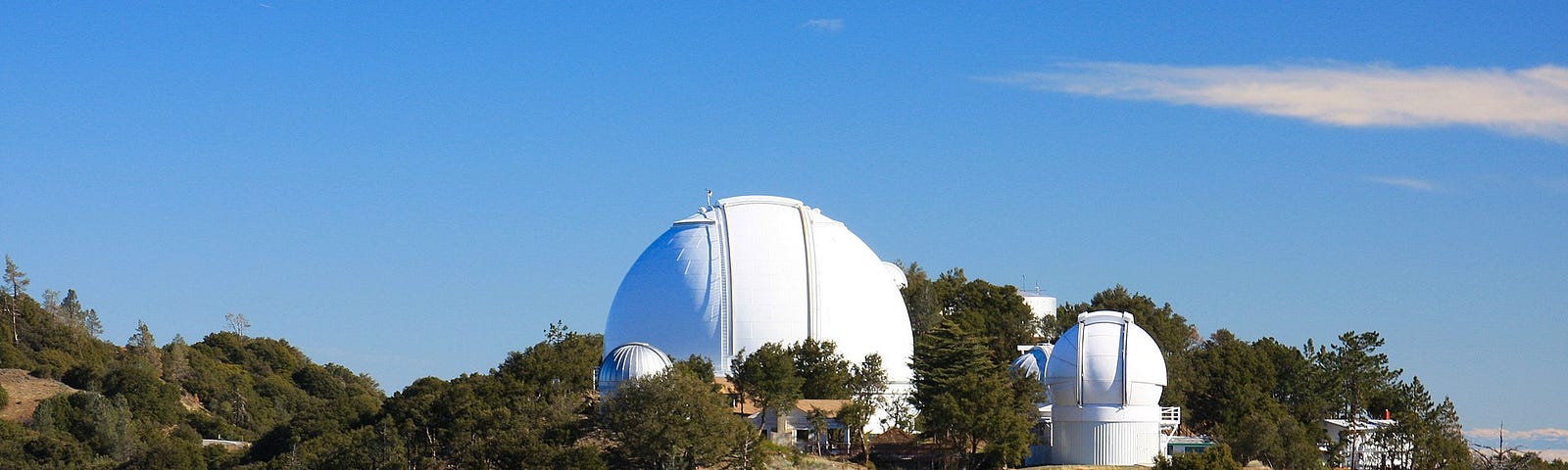 Photo of Lick Observatory’s 120-inch Shane telescope on Mt Hamilton above East San José, CA. Skies are a brilliant blue, with one cloud to the right, with another smaller telescope to the right, and a building in the foreground. Trees and foliage surround the telescopes on a hill with a driveway leading to them.