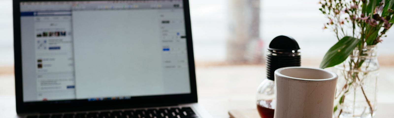 A picture of an open laptop, a mug, flowers in a vase and an open book with a pen on it on top of a desk.