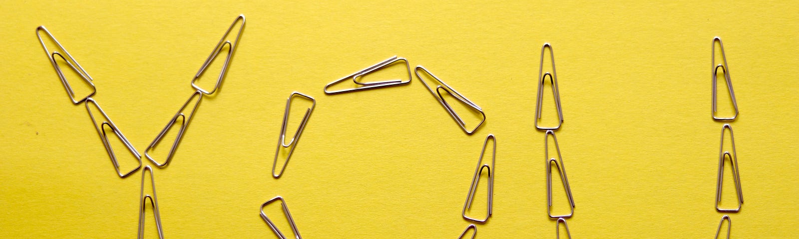 The word “YOU” portrayed by brass paperclips on a yellow matte.