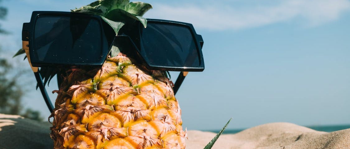 A pineapple wearing sunglasses, resting in sand on a beach