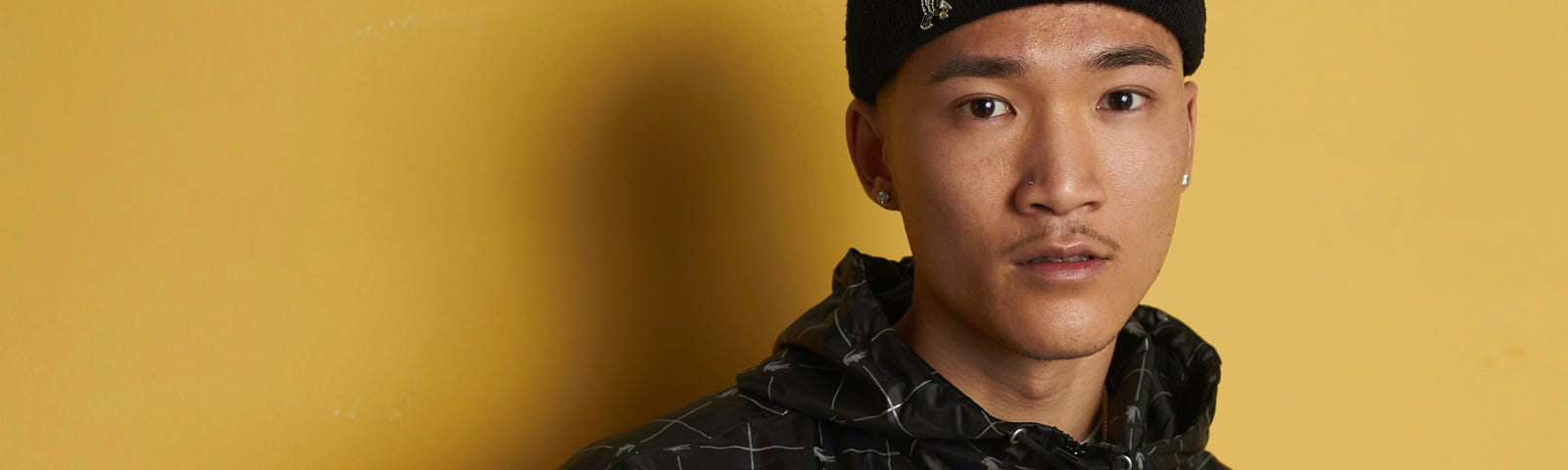 An Asian young man wearing all black is propped against a yellow wall.