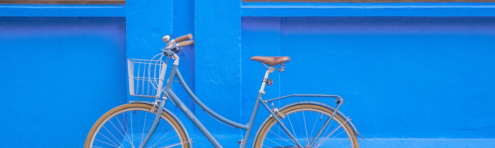 Basic bicycle with a basket on the front, on a footpath, leaning against a bright blue wall.