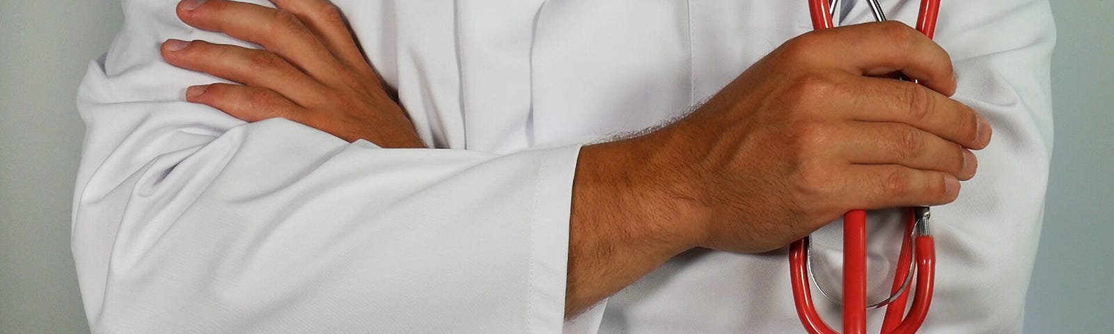 Close-up of arms crossed over in front of a body wearing a white lab coat and holding a stethoscope in one hand.