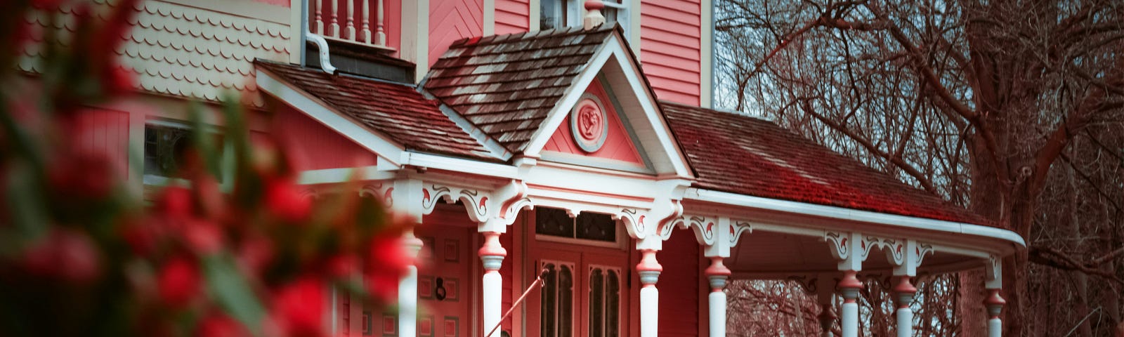 Pink Victorian home with large porch and red bushes.