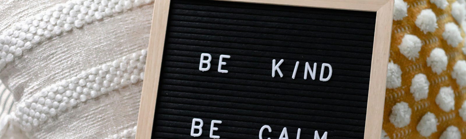 Board that says BE KIND BE CALM BE SAFE #SPREADJOY leaning against cream-colored and yellow pillows.