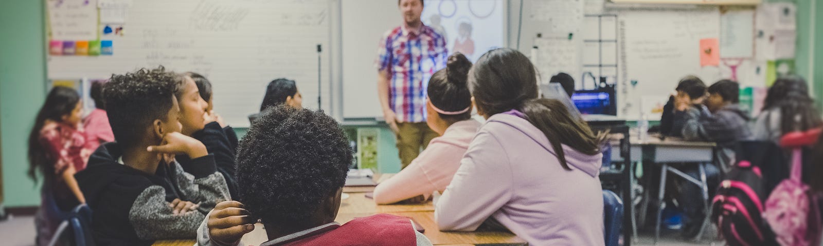 Middle-school-age students in a classroom listening to a male teacher in a plaid shirt