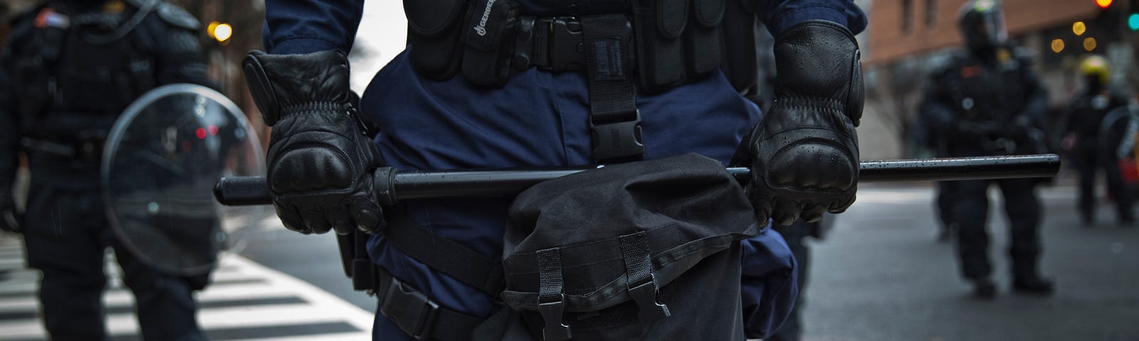 A close-up view of one riot policeman from chest down with long black baton held menacingly across his thighs. He is flanked in the distance with other similarly-clad officers with shields and helmets.