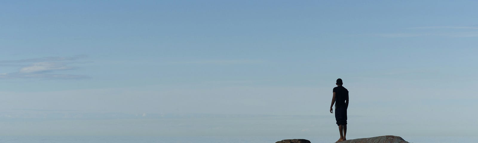A man standing on a large boulder in the middle of a still ocean, staring at the horizon, where blue sky meets blue sea.