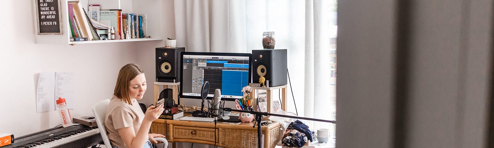 A woman sits at her desk in her bedroom. She is on her phone. On her desk is her computer. Behind her is a keyboard, next to her is recording equipment. To the far right is her bed and on the walls are shelves filled with books.