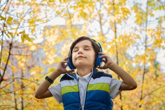 Young boy in striped jacket holding his headphones with chin raised and eyes closed with yellow-leaved trees in background