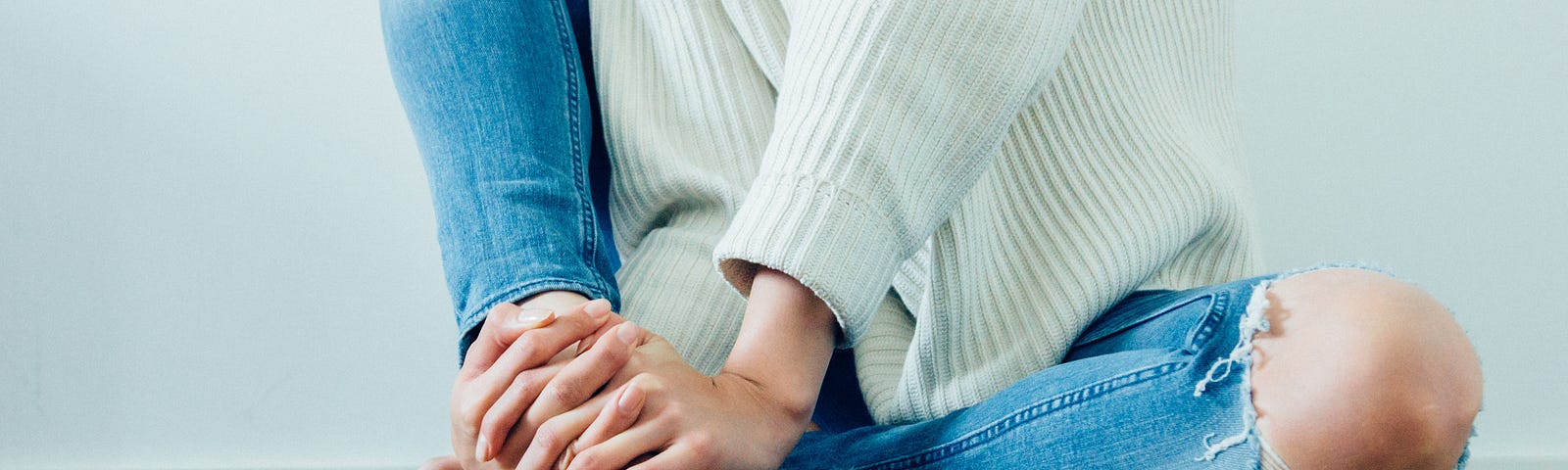 A woman sits on the floor wearing jeans and a white jumper. One knee is up and her hands are clasped on her foot.